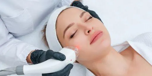 A person receiving a facial laser treatment with a professional using a handheld device, both wearing protective gear, in a clinical setting.