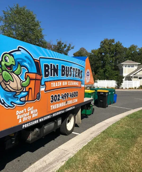 A Bin Busters trash bin cleaning truck with a turtle mascot, parked next to several trash bins.