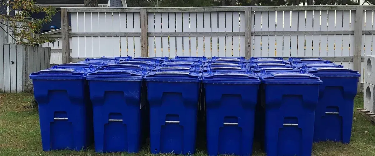 Multiple rows of blue recycling bins with closed lids sit on green grass in front of a white wooden fence.