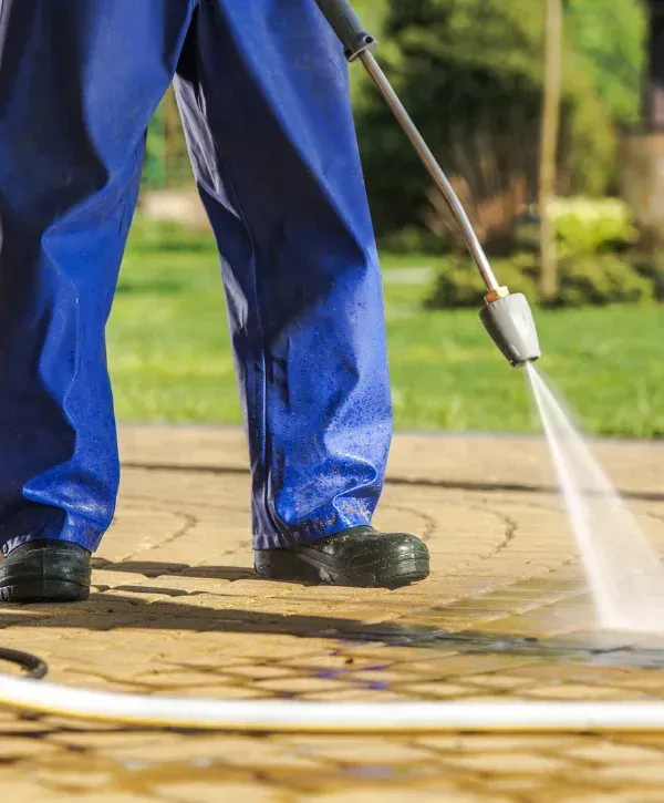 A person in blue protective trousers and boots pressure washing a dirty paved driveway on a sunny day.