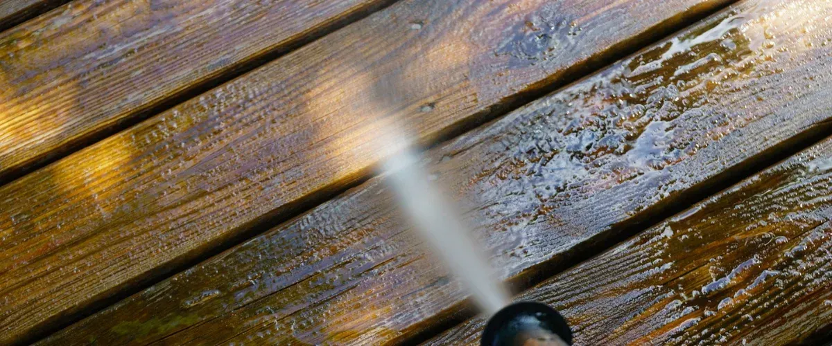 A close-up view of a wooden deck being power-washed, with a stream of water cleaning the planks.
