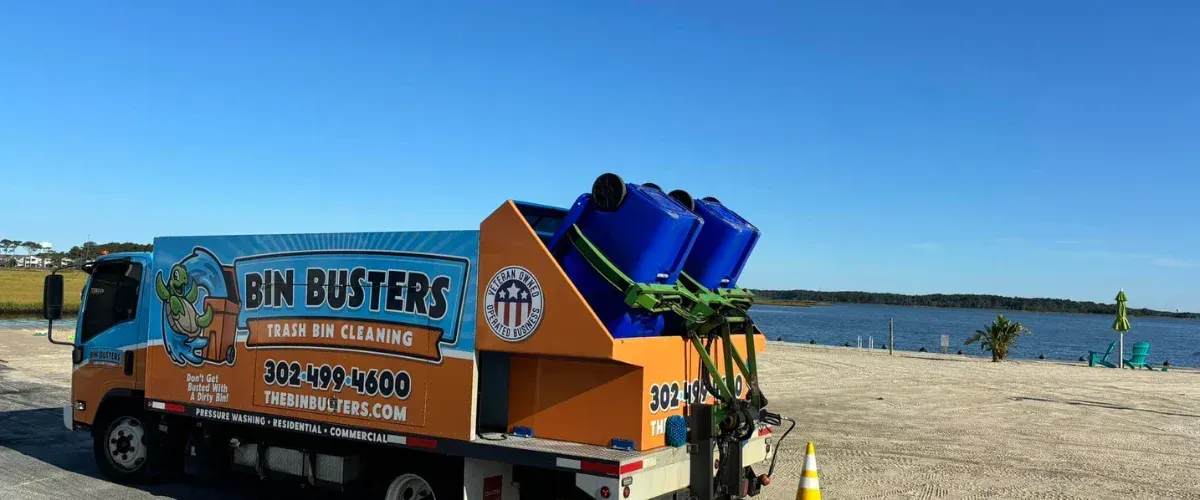 A Bin Busters trash bin cleaning truck with blue bins on its rear, parked on a sandy waterfront with water and trees in the background.