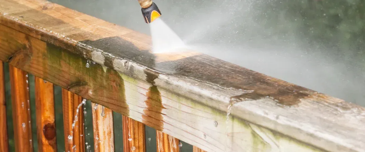 A yellow pressure washer nozzle sprays a strong stream of water, cleaning dirt off a concrete path with green grass nearby.