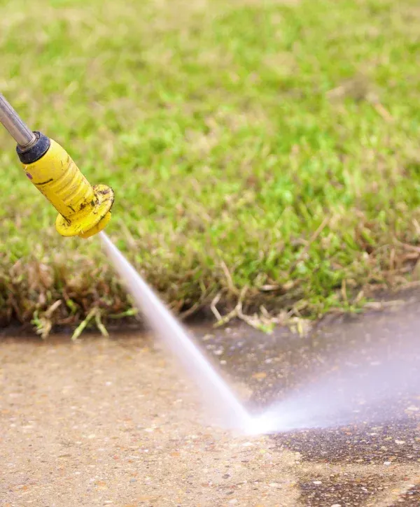 A yellow pressure washer nozzle sprays a strong stream of water, cleaning dirt off a concrete path with green grass nearby.