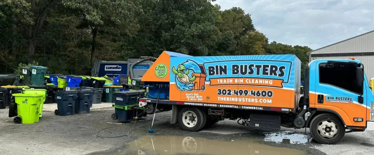 A blue and orange Bin Busters trash bin cleaning truck with a turtle mascot, parked on a gravel lot next to multiple bins.
