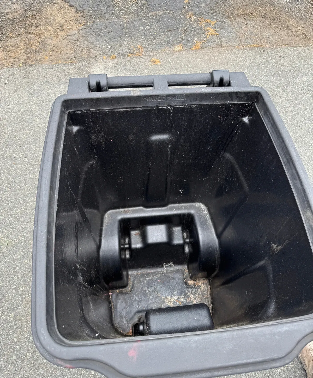 An overhead view looking down into an empty, dirty black plastic wheeled trash can with 'RUGGED RIM' embossed inside.