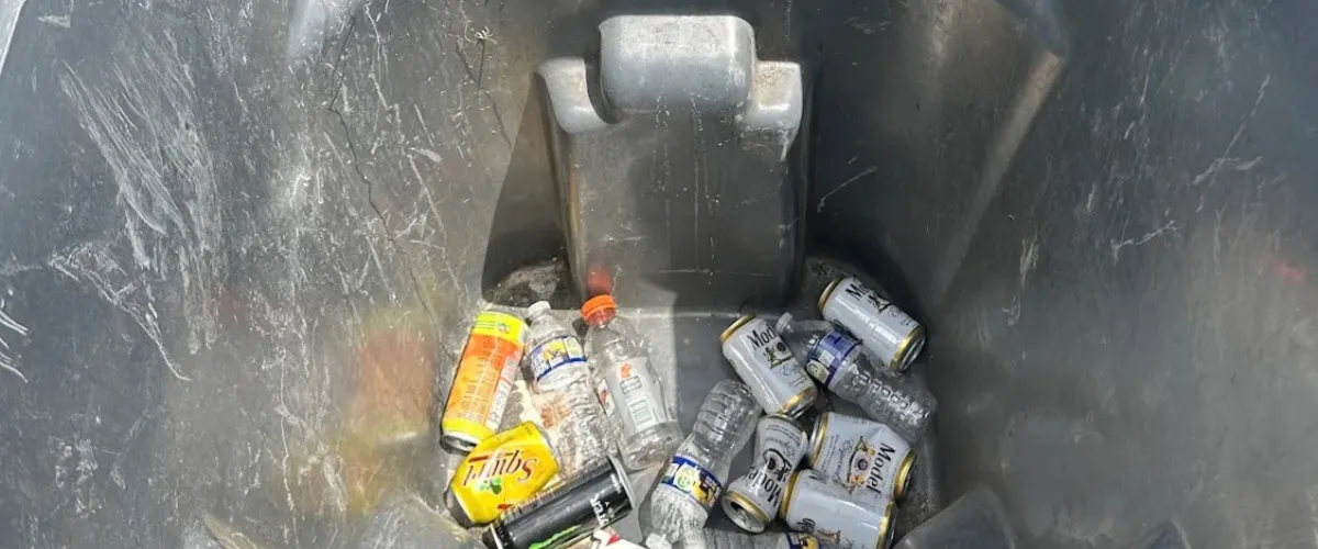 An overhead view of a gray recycling bin filled with various empty aluminum cans and plastic bottles.