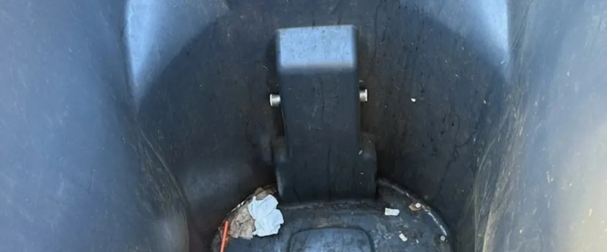 A top-down view inside a dark, dirty plastic trash bin showing a structural support and small pieces of trash at the bottom.