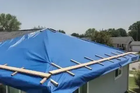 Blue emergency tarp secured with wood strips over a storm-damaged roof in Blaine