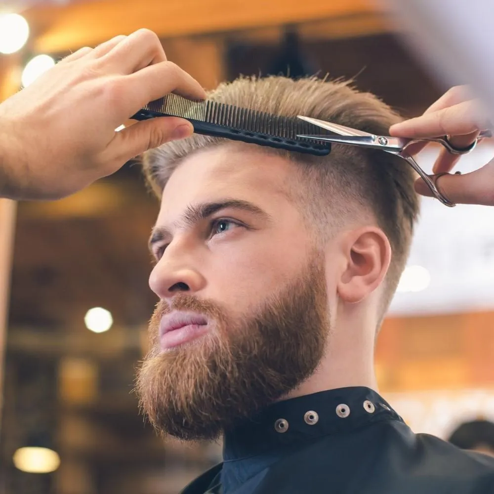 Professional barber styling a client's pompadour haircut and full beard with shears and a comb at a family barbershop.