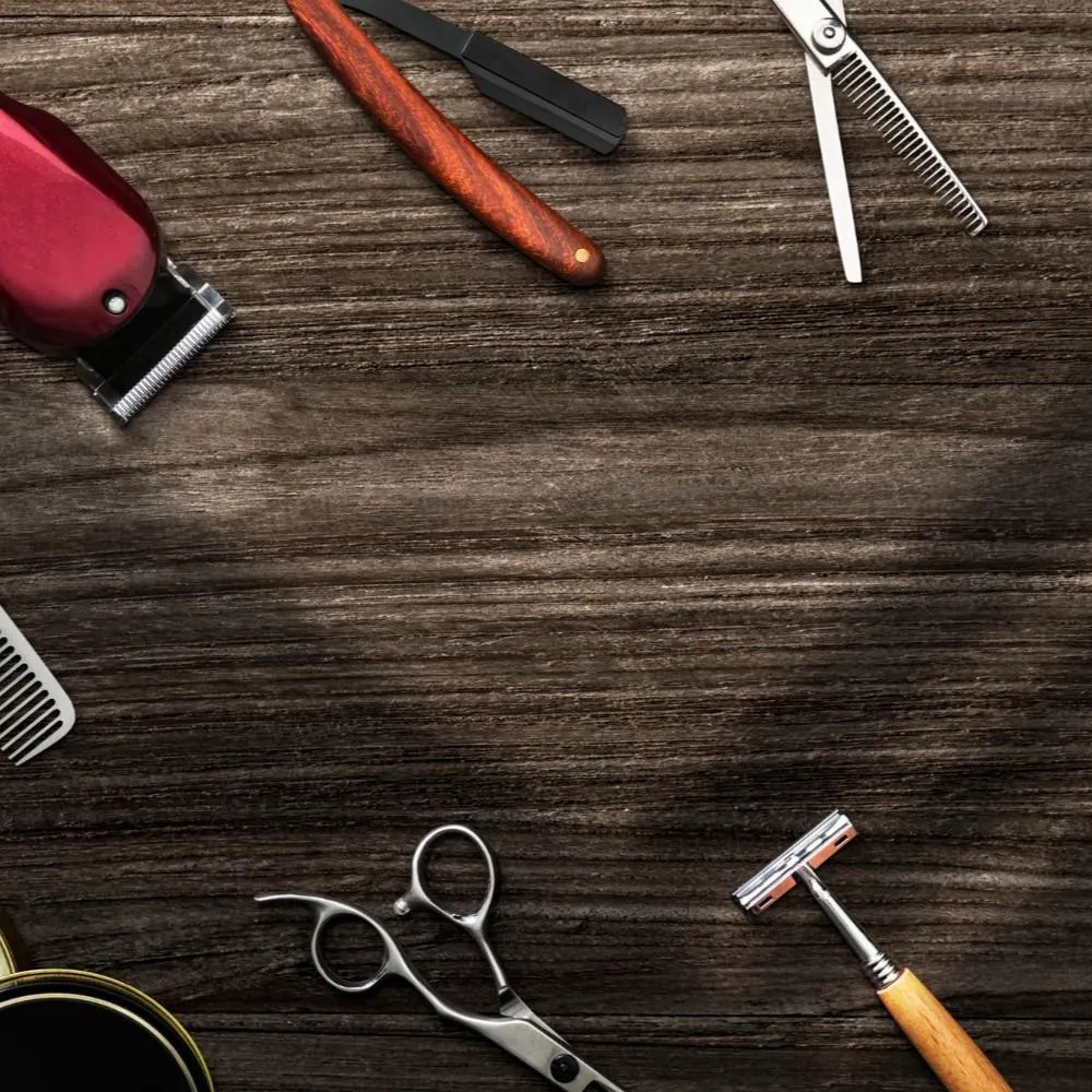 Flat lay of professional barber tools including hair clippers, shears, a straight razor, and a comb arranged on a dark wood background.