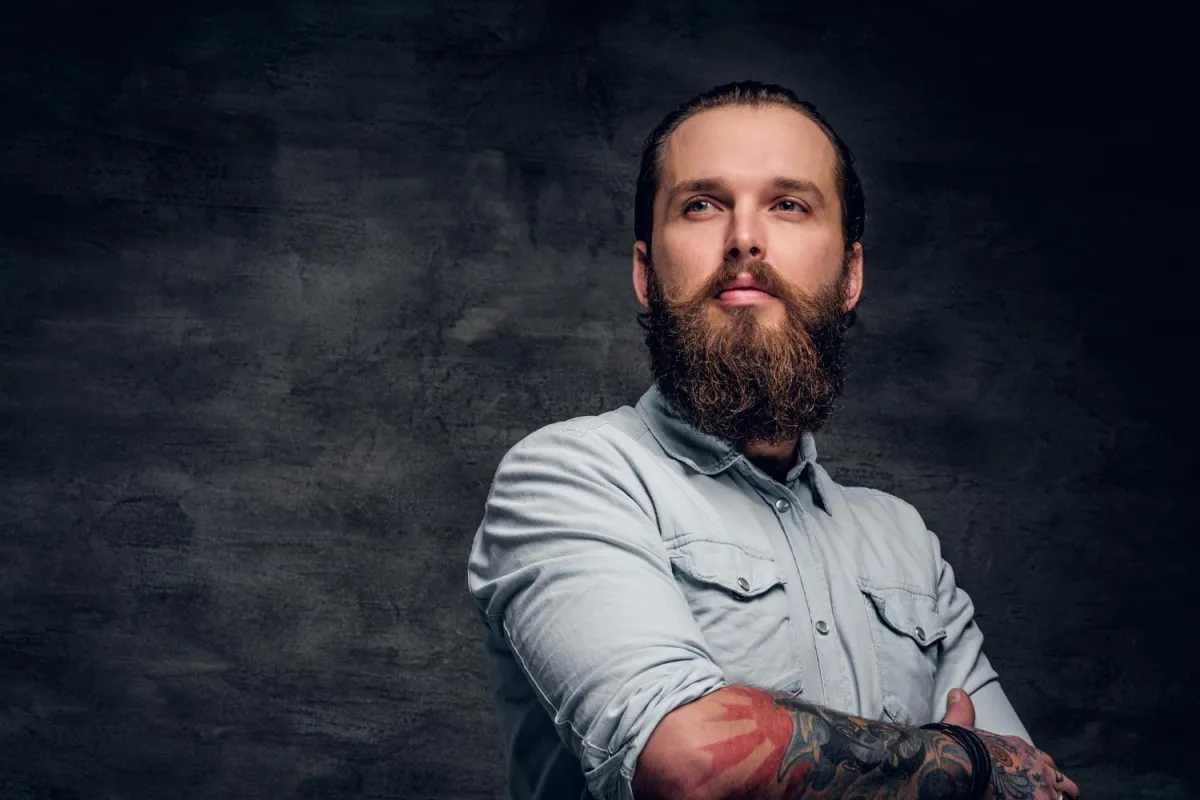 Man with a perfectly groomed long beard and mustache posing against a dark textured background after a professional beard trim.
