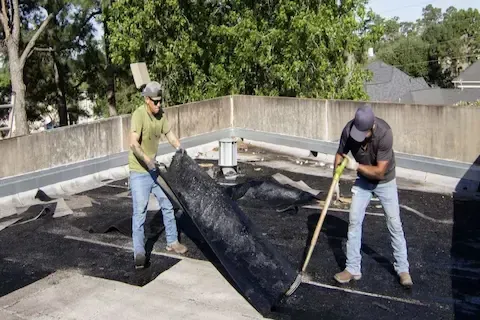 Two workers peeling back old asphalt roofing material during flat roof replacement in Plymouth
