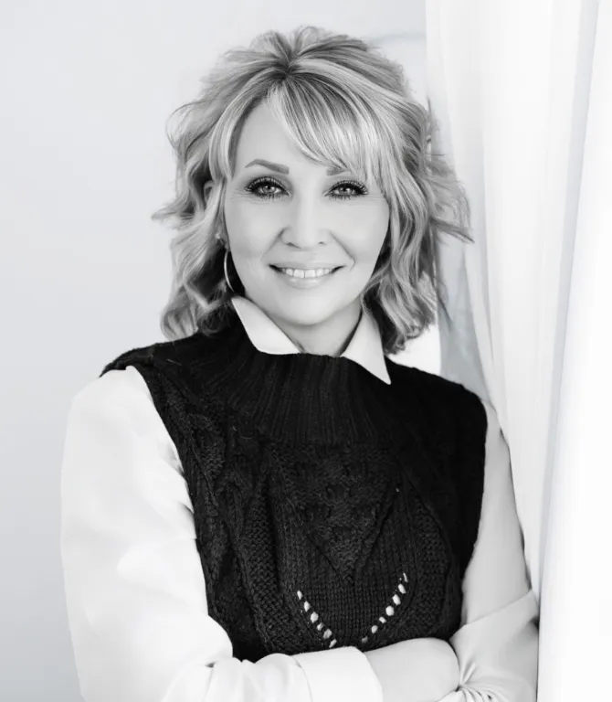 Black and white portrait of a blonde woman wearing a dark patterned top, smiling warmly in a bright setting.