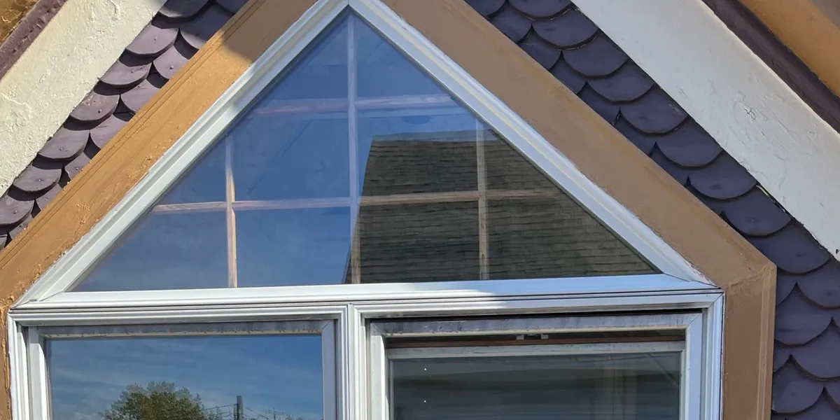 Exterior view of a residential window with triangular upper glass, white window frames, and decorative siding.