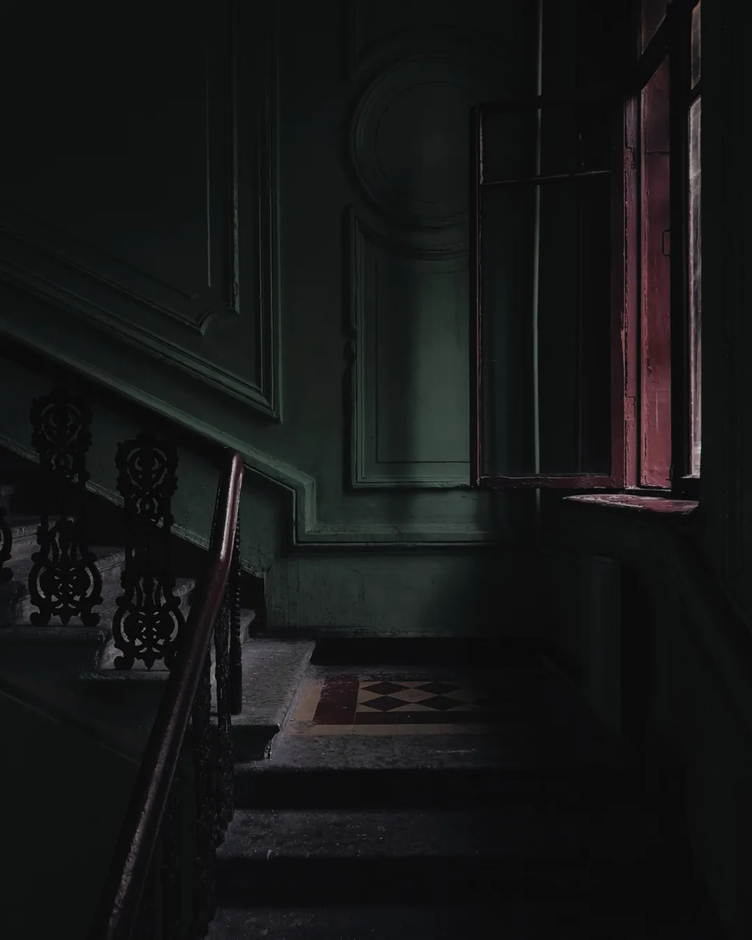A dark image of a staircase, there is a window to the right, in which the wall looks to be painted deep red. The light is low, and thee are shadows across the image
