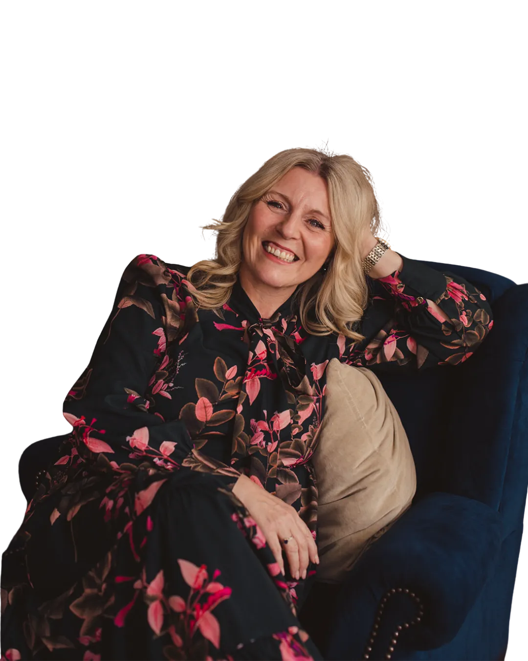 Jacqueline is a blonde woman with smooth elegant curly hair. She is wearing a black dress adorned with pink and deep green florals. She is sat on a dark blue armchair with one elbow resting on the back of the chair and her head resting on her hand. Her silver watch is visible on her wrist. She looks happy, smiling at the camera as though she is glad that you're here. 