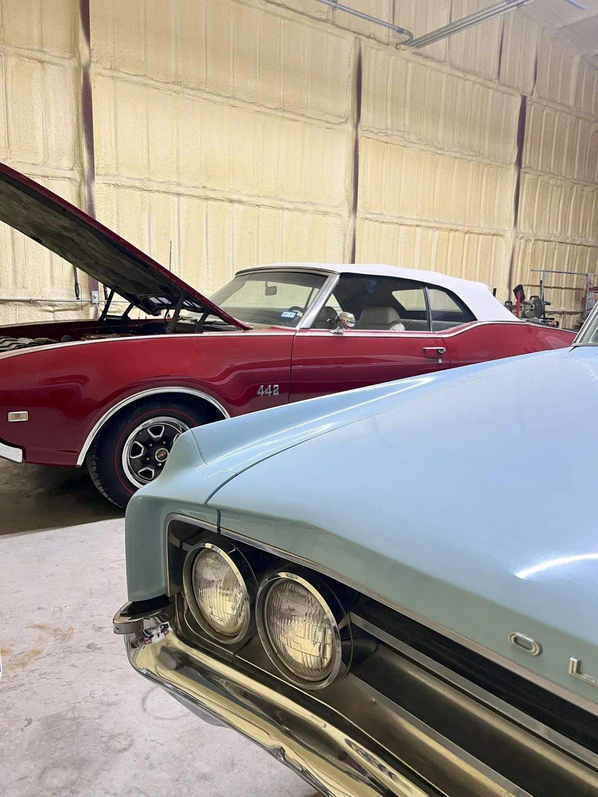 Vintage cars in a garage, featuring a Oldsmobile 442 red convertible with the hood raised and a light blue vehicle in the foreground, showcasing Kanewske Motors' automotive restoration services.