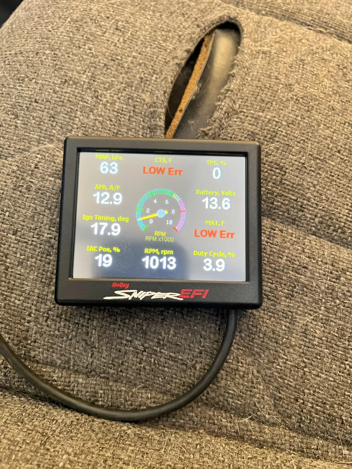 Display screen of Holley Sniper EFI showing engine diagnostics, including RPM, air-fuel ratio, and error messages, on a textured fabric background.