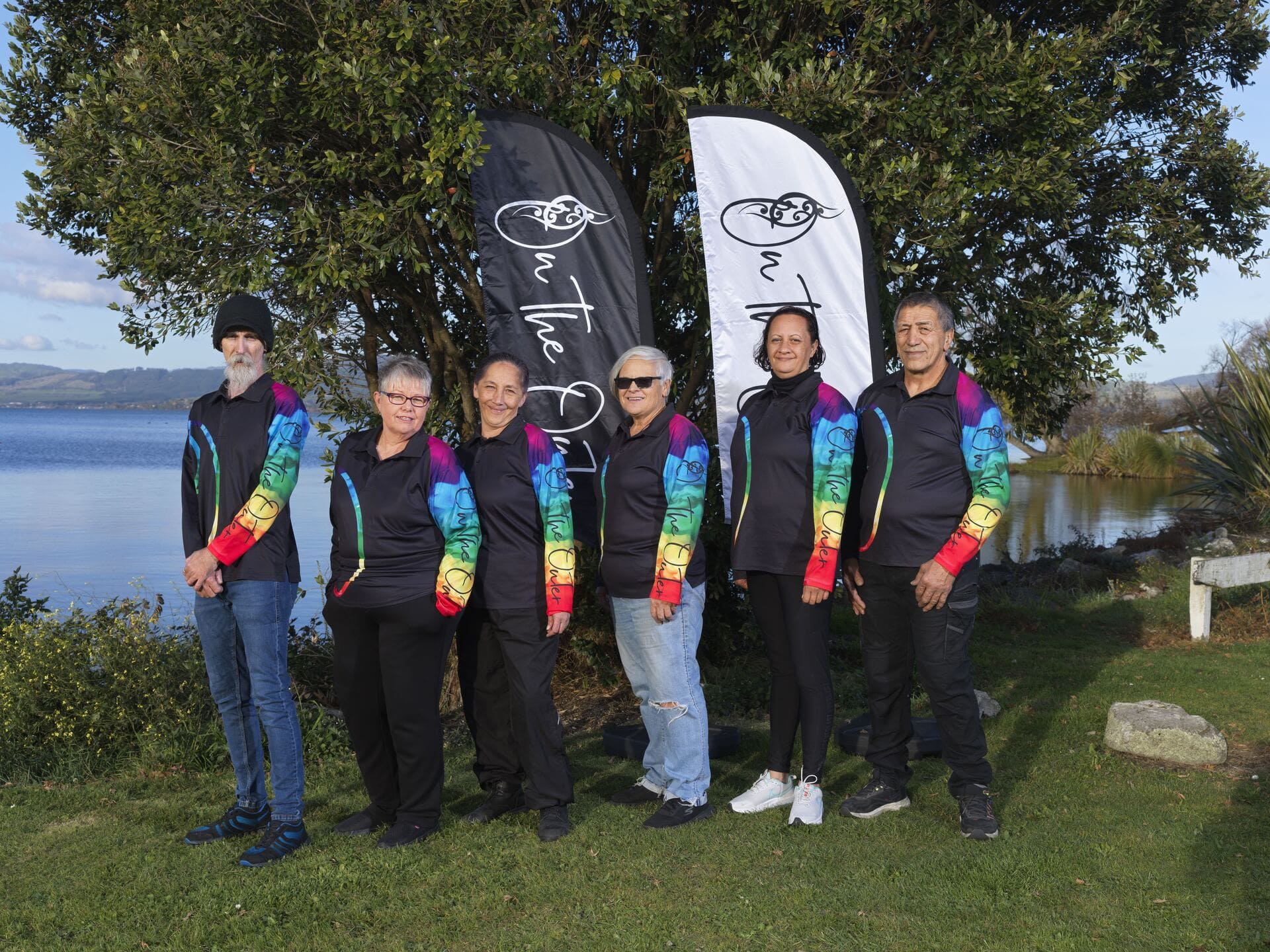 Six people wearing matching black tops with rainbow-coloured sleeves beside On the Outer flags