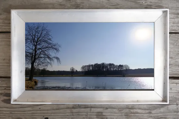 Gerahmtes Landschaftsbild mit See, Bäumen und Frühlingssonne – ruhiger Moment einer Schwellenzeit zwischen Winter und Frühling.