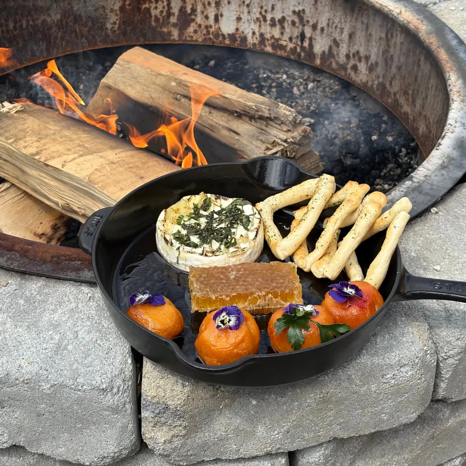 An outdoor fire pit culinary experience featuring baked brie, honeycomb, and crackers served warm in a cast-iron skillet.