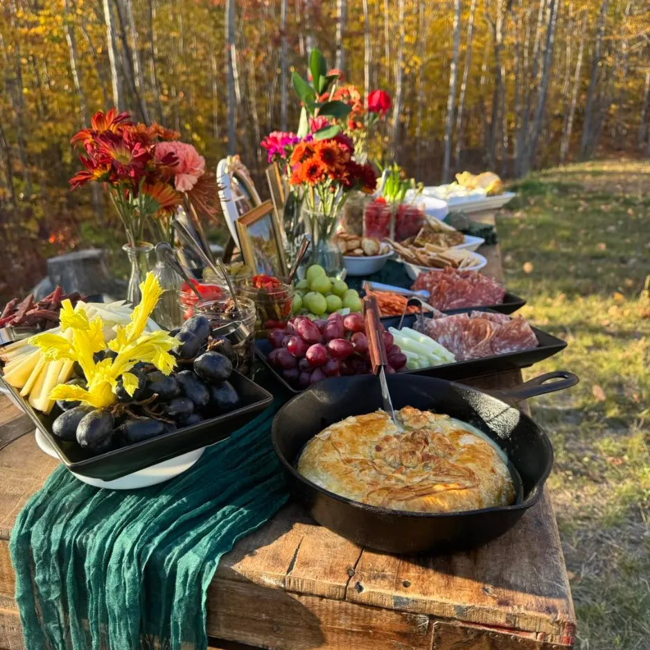 An outdoor autumn buffet table with floral arrangements, fresh fruits, and hot dishes overlooking a scenic Maine forest backdrop.