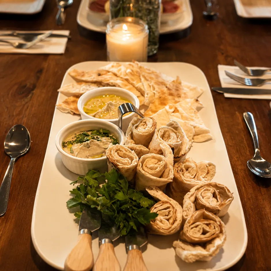 An indoor dining setup with a candlelit table and a platter of rolled flatbreads, dips, and fresh herbs.