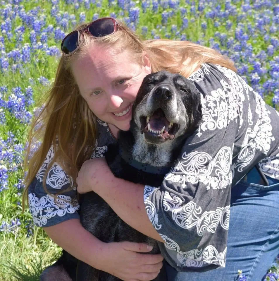 Dog trainer Brandie Sims with older black lab dog sitting in a field of Texas bluebonnets