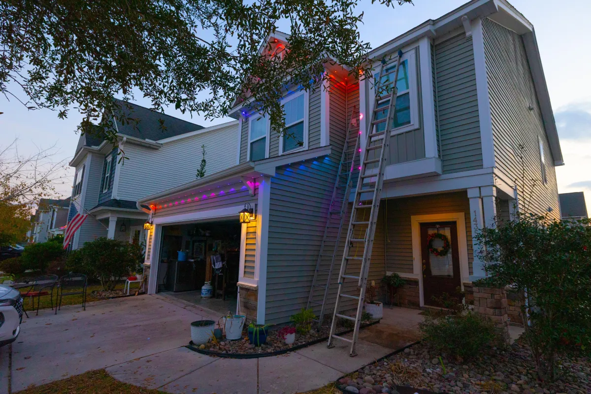 Two-story home with multicolor permanent LED outdoor lights installed along the roofline, featuring a ladder and open garage at dusk.