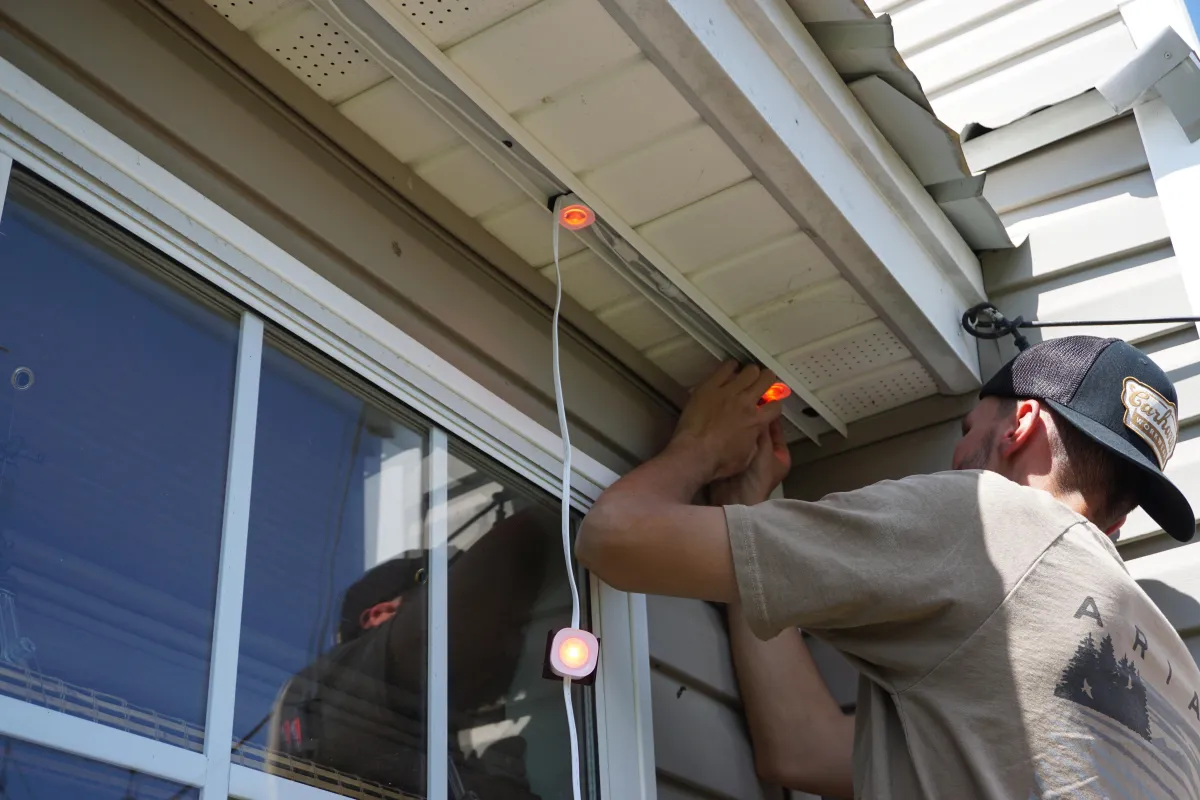 Two-story home with multicolor permanent LED outdoor lights installed along the roofline, featuring a ladder and open garage at dusk.