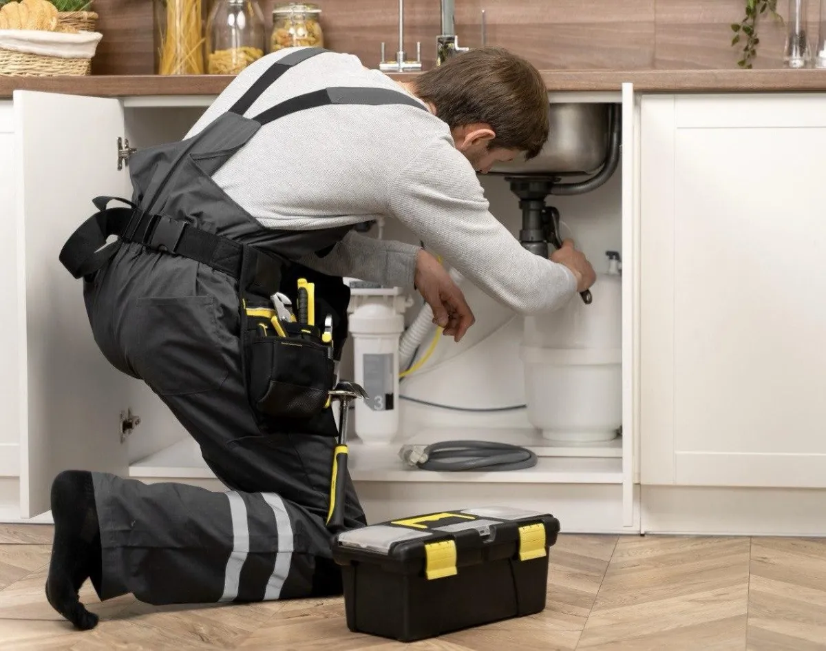 Plumber kneeling under a kitchen sink repairing drain pipes