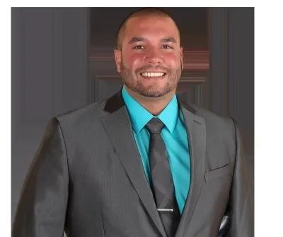 Portrait of a smiling female real estate agent in her 30s, African American, wearing a navy blazer and white blouse, standing in front of a Portage home with a welcoming expression. 1:1 aspect ratio.