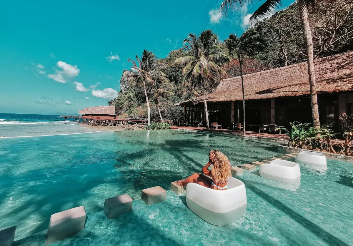 Woman relaxing on a floating chair in a luxurious infinity pool overlooking the ocean, surrounded by tropical palm trees and a thatched-roof resort in a scenic island paradise.