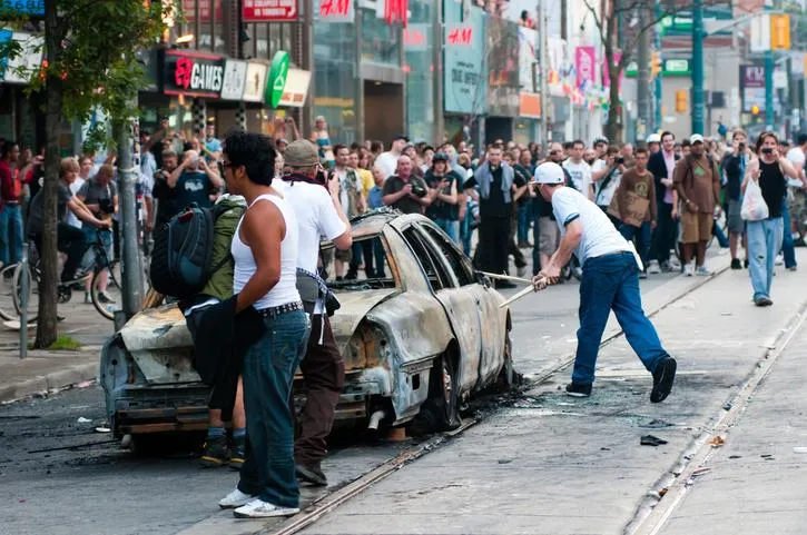 Rioters standing around the remains of a burnt vehicle in a riot.