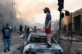 A rioter standing on the hood of a burned vehicle in the middle of a riot.