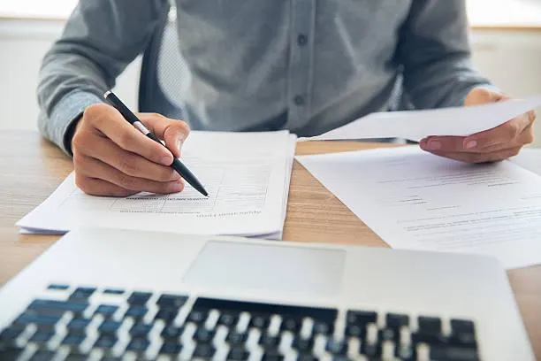 Person reviewing documents and filling out forms for insurance claims with a laptop in the background