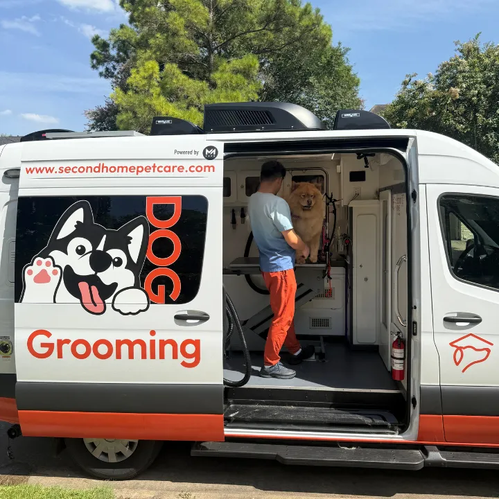 Professional mobile pet grooming van parked in Katy, TX, with a groomer gently styling a fluffy dog inside, presented on a warm peach background with playful paw print and heart accents.