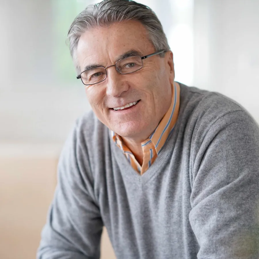Smiling older man wearing glasses and grey sweater, posing indoors in natural light