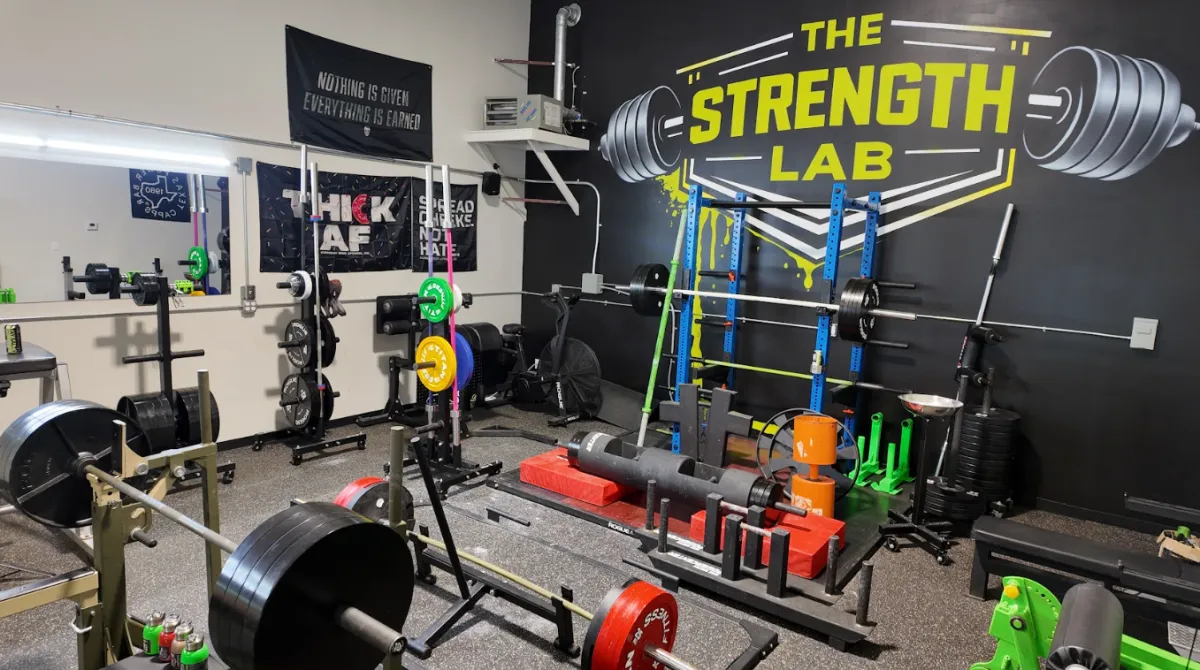 Extensive interior view of The Strength Lab gym in Newtown, Connecticut, a premier training facility for powerlifting and strongman athletes. The image captures custom squat racks, weight plate trees, calibrated kilo plates, deadlift platforms with strongman implements, and a large black and yellow branded wall mural. Motivational flags are visible on the wall.
