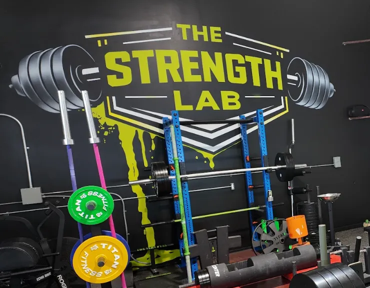 he Strength Lab branded logo wall mural in toxic yellow and black. The scene showcases a blue powerlifting squat rack with a loaded barbell, specialized barbells on a vertical stand, vibrant yellow and green Titan Fitness weight plates, and a heavy strongman log training implement in Newtown, Connecticut.