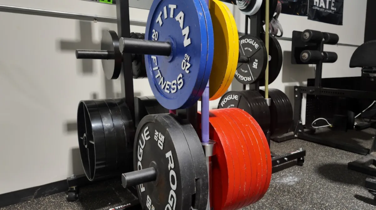 Close-up of competition weight plate storage trees at The Strength Lab near Danbury, CT. Pictured are tournament-grade black Rogue calibrated kilo plates, vibrant blue and yellow Titan Fitness weight plates, and a vertical barbell storage stand on a premium rubber gym floor.