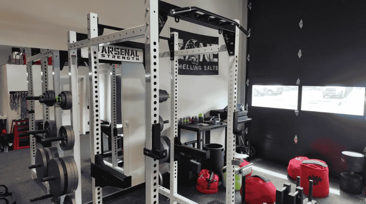 Heavy-duty white Arsenal Strength power racks with custom multi-grip pull-up bars stationed next to heavy strongman training sandbags and an open garage bay door inside a 24/7 access gym near Shelton, CT.