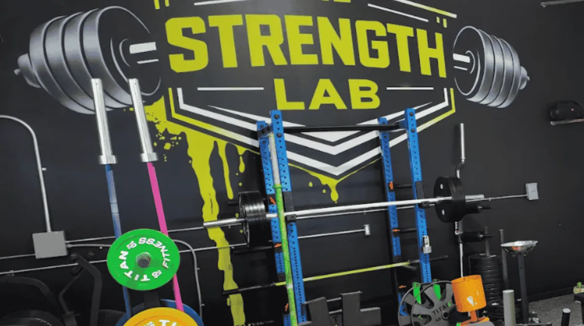 The Strength Lab signature toxic yellow and black wall mural logo in Newtown, CT. The close-up view highlights a blue steel powerlifting rack with a loaded barbell, specialized barbells in a vertical storage tree, and bright green and yellow Titan Fitness weight plates.