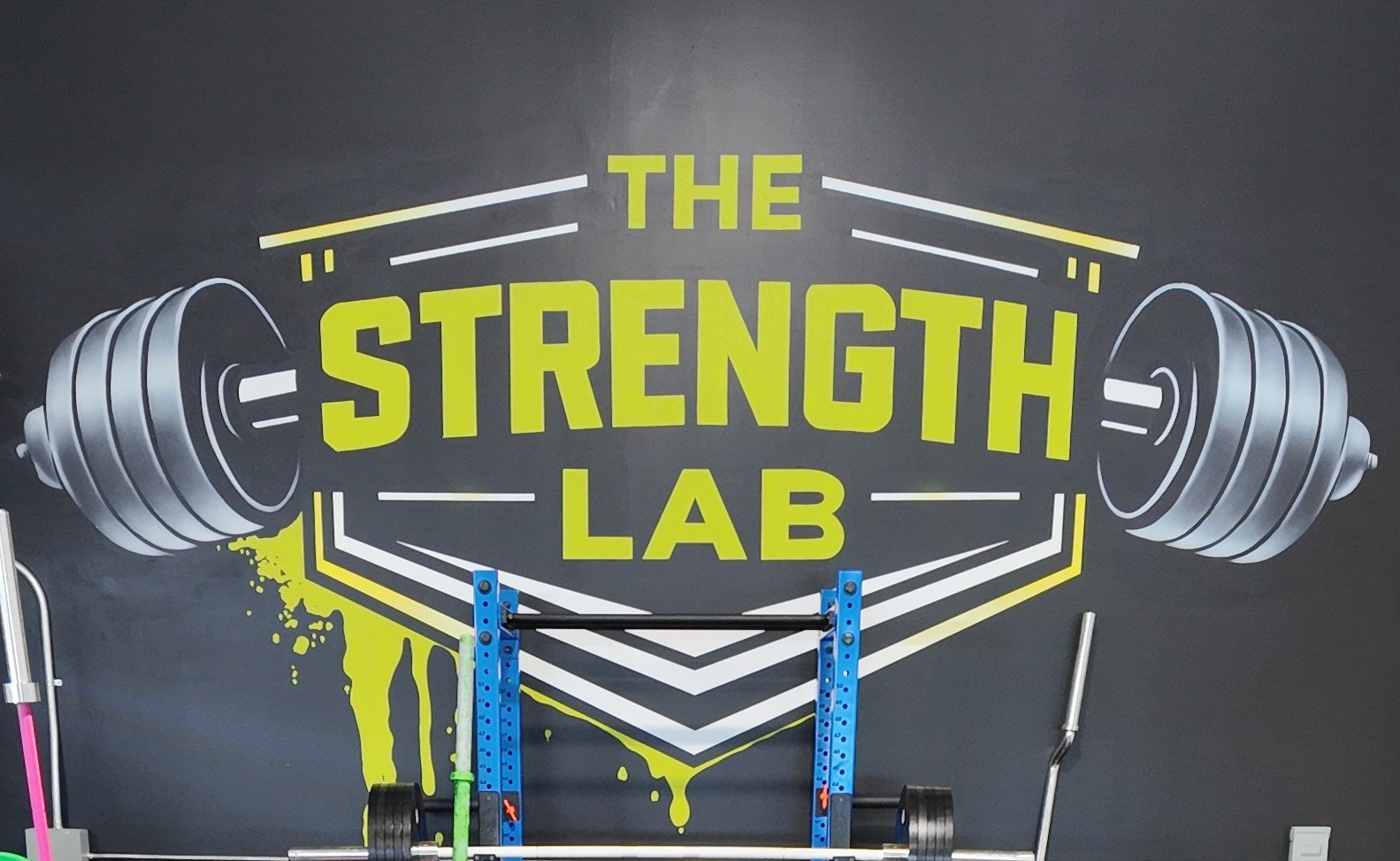 Symmetrical front shot of the custom black and yellow logo mural inside The Strength Lab in Newtown, Connecticut. A blue steel squat rack loaded with plates sits directly under the branding, surrounded by professional powerlifting and strongman gear.
