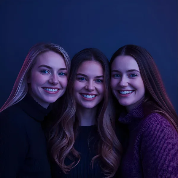 Portrait of three women standing close together and smiling, illuminated with cool blue and purple tones on a dark background.