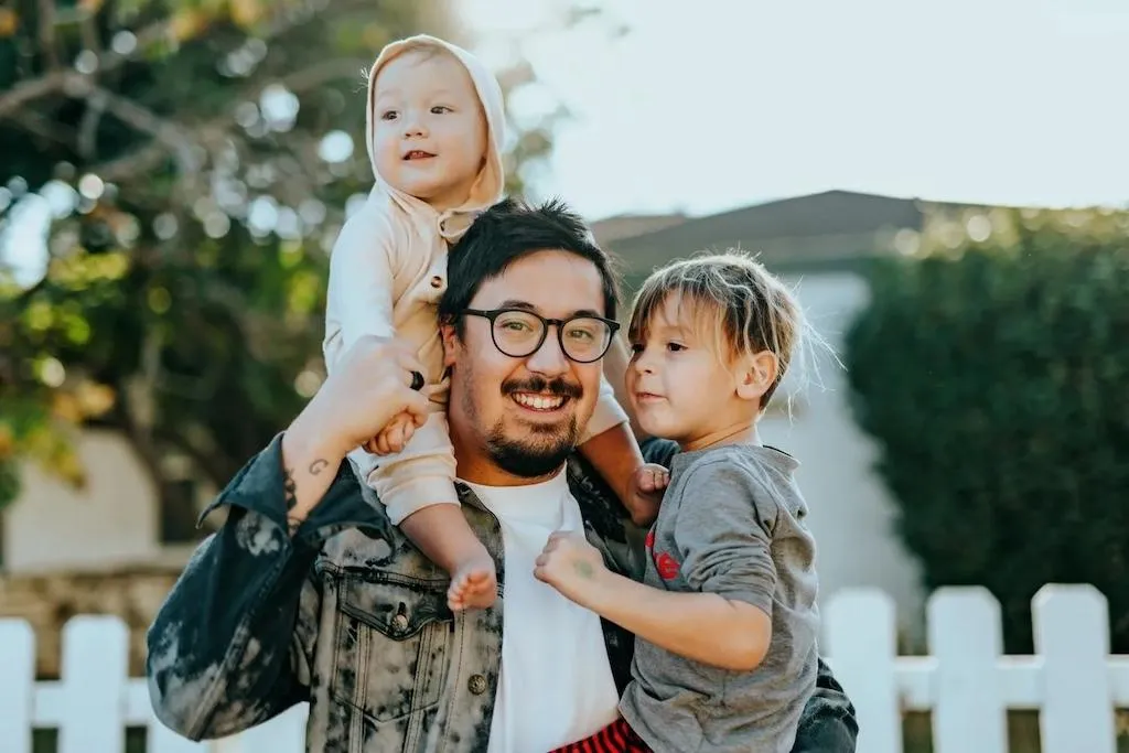 A man wearing glasses and a child pose in front of a white picket fence, representing the dream of homeownership.