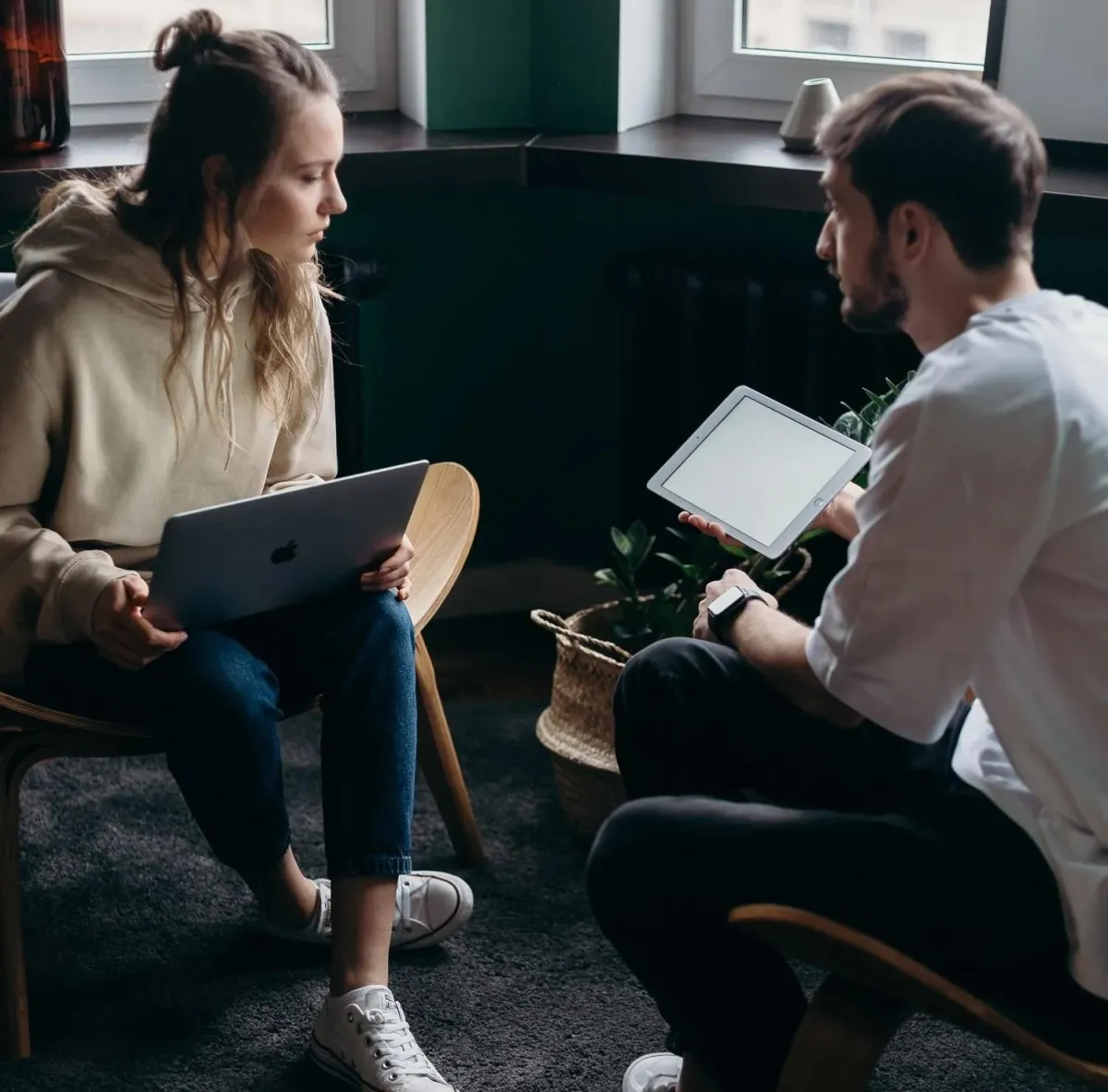 Two people sit indoors discussing something. One holds a laptop, and the other holds a tablet. They appear focused, sitting in a modern room with large windows.