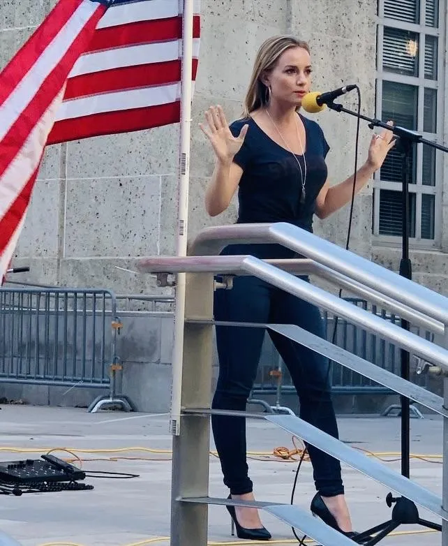 ivory hecker speaking on microphone beside the american flag