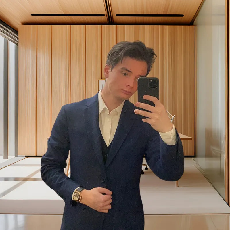 A middle-aged man with glasses and a salt-and-pepper beard, wearing a crisp white shirt, sits at a desk with architectural plans. The background features large windows and city views, suggesting an urban office. The image conveys expertise and thoughtful focus.